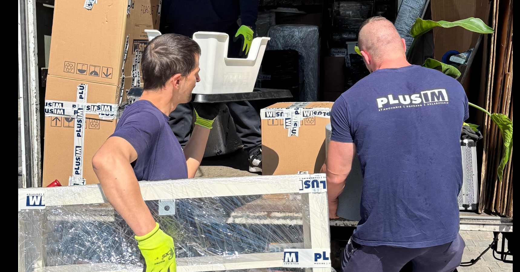 Pracovníci spoločnosti PLUSIM profesionálne manipulujú s nábytkom a balenými krabicami pri sťahovaní. Bezpečné nakladanie vybavenia do nákladného vozidla s použitím ochranných pomôcok a špeciálneho obalového materiálu, typické pre komplexné sťahovacie